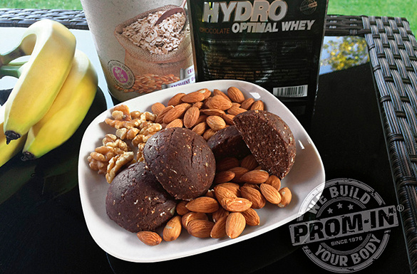 Lifestyle fotografia s čiernou dózou Hydro Optimal Whey na ratanovom stole. Na bielom tanieri sú čokoládové sušienky, mandle a vlašské orechy. V pozadí banány a pohár s čokoládovým nápojom. Dole vpravo je logo Prom-in "Build Your Body". Celá scéna je vonku na záhrade s rozmazaným zeleným pozadím.