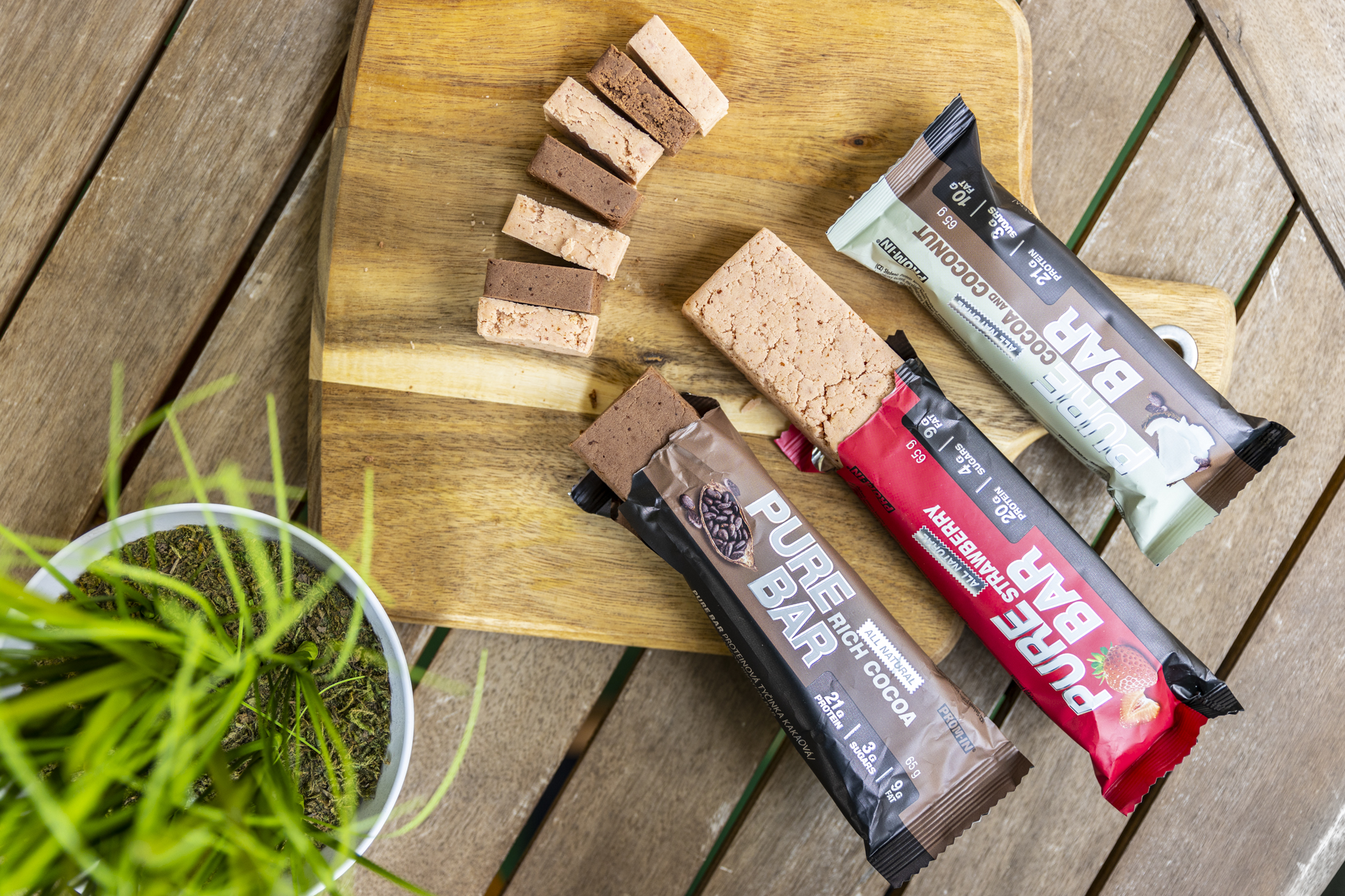 Na drevenej doske sú rozložené tri proteinové tyčinky Pure Bar - hnedá s čokoládovou príchuťou, červená s jahodovou príchuťou a bielo-hnedá s kokosovou príchuťou. Vedľa tyčiniek sú nakrájané kúsky tyčiniek ukazujúce ich vnútornú štruktúru a textúru. V ľavom dolnom rohu je biely kvetináč so zelenou rastlinou. Celá scéna je na drevenom terasovom podklade.