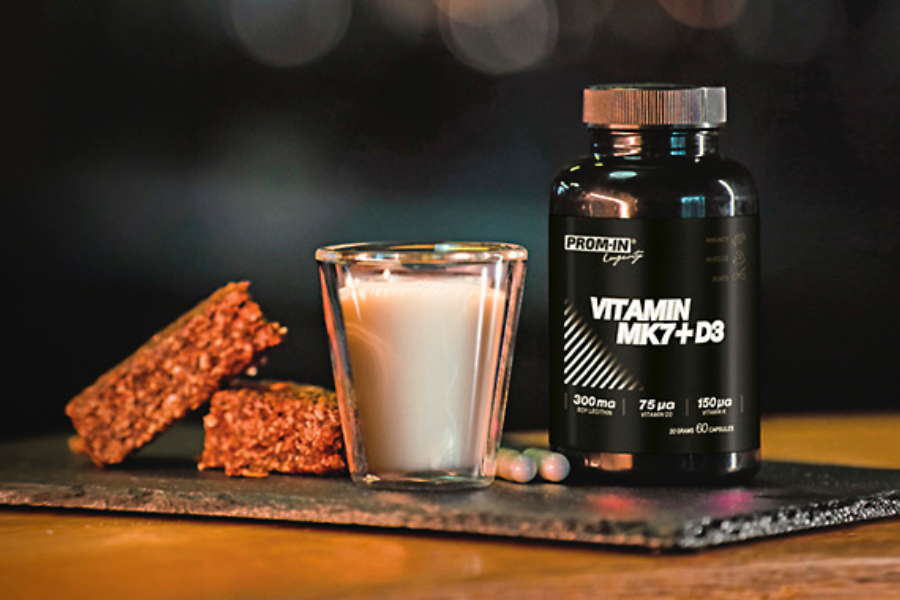 Ilustračný obrázok vitamínu MK7 + D3. Čierna fľaštička produktu Vitamín MK7 + D3 s drinkom na stole.