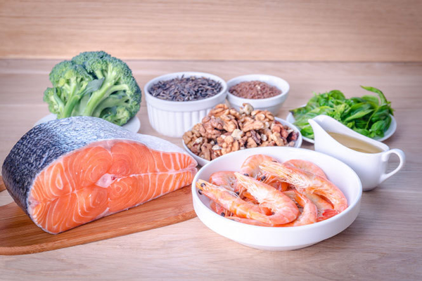 Ilustračná fotografia potravín, v ktorých je vysoký obsah omega 3 mastných kyselín. Losos, krevety, orechy, brokolica, špenát a semienka odprezentované na tanieroch a v miskách na stole.